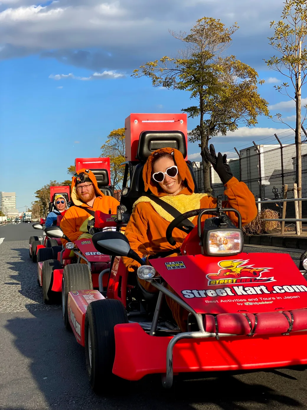 People in orange animal-costume outfits driving red Street Kart go-karts down a city street on a sunny day.
