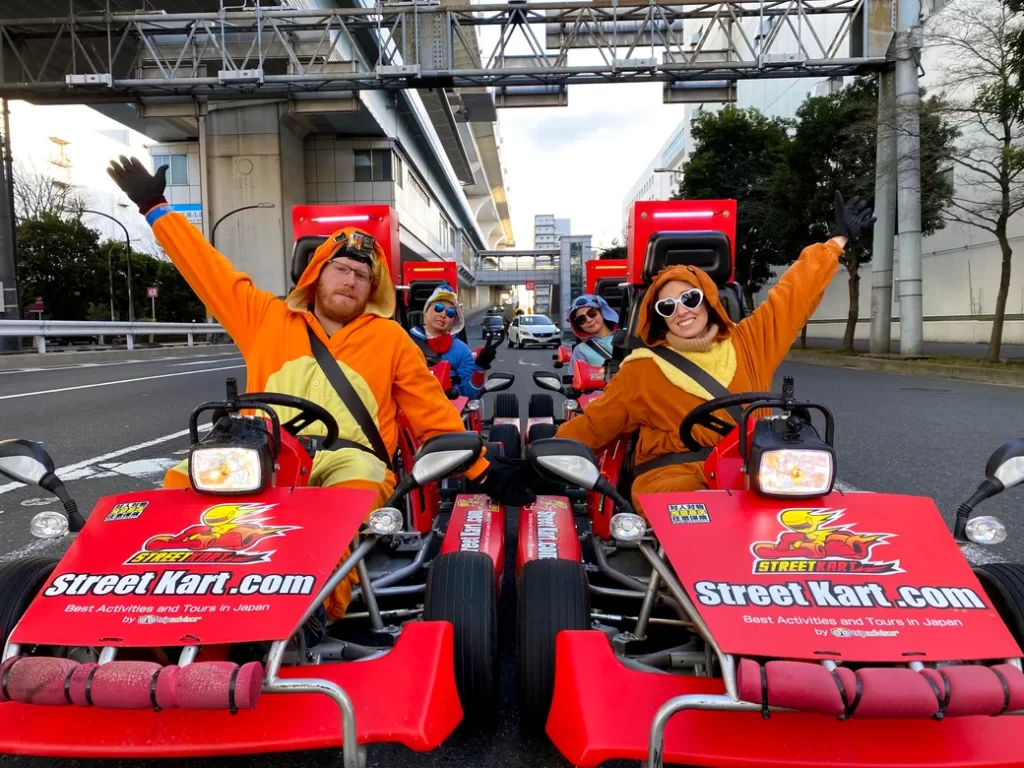 IMG_3612 - StreetKart Four people in bright orange mascot outfits ride red Street Kart go-karts on a city street, waving their arms at the camera under an elevated track.