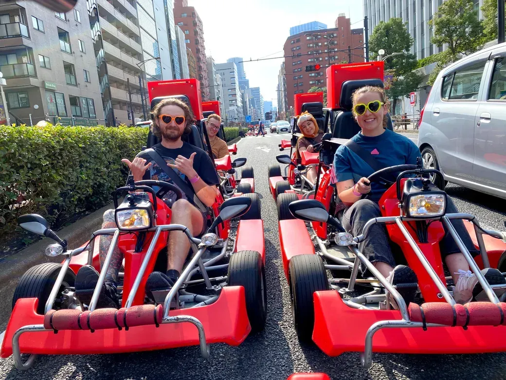 Group of friends riding red pedal go-karts on a city street, wearing sunglasses and smiling.